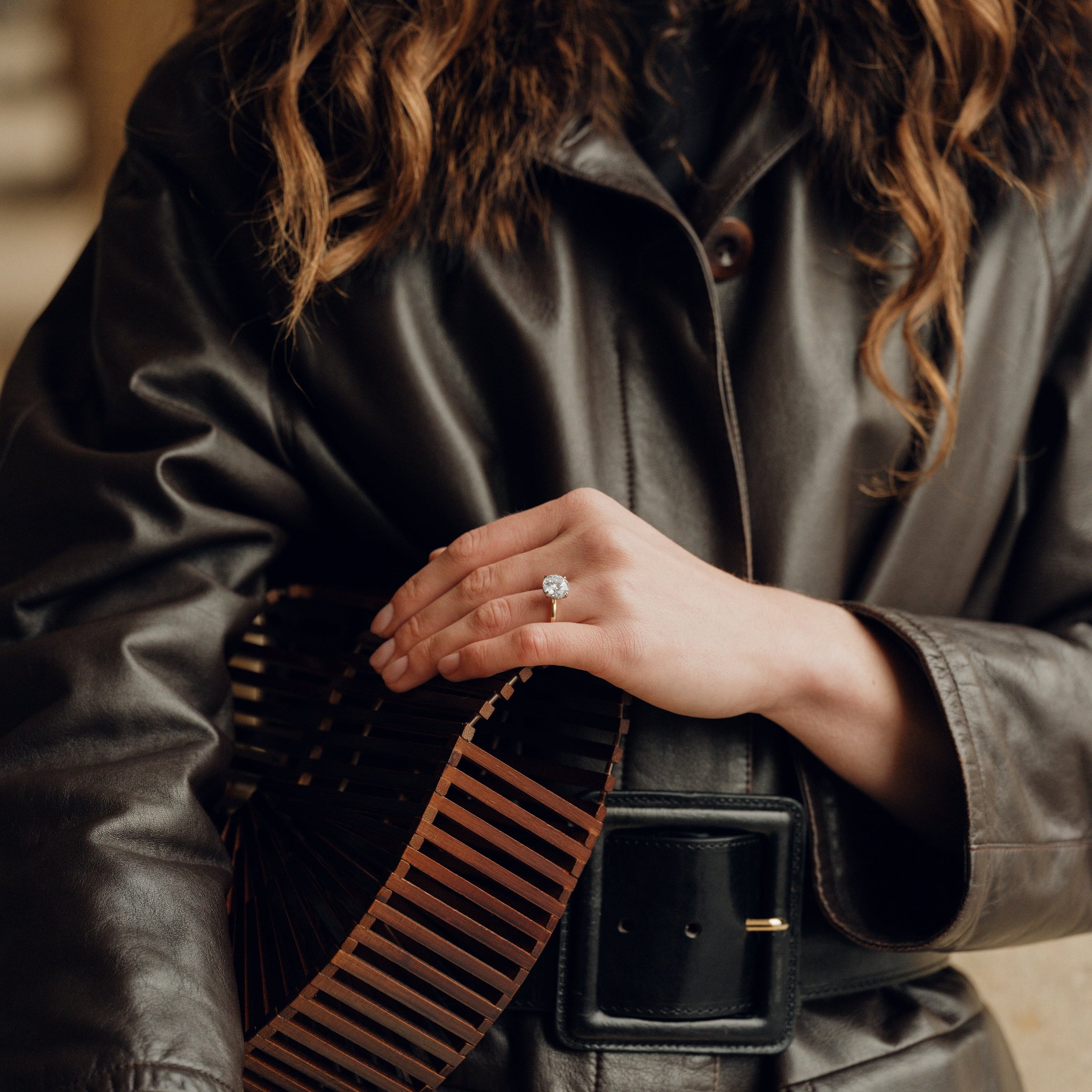 Person wearing a dark leather jacket with a fur-lined hood, holding a brown and black striped bag.